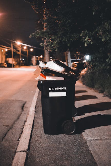 On a pavement near a street corner, there is a small pile of rubbish consisting of a black bag, a cardboard box, and a blue and white cardboard container, possibly for household waste or packaging mat