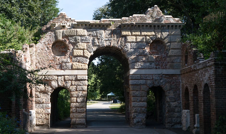 A large historic stone archway stands as the central feature of this outdoor scene, constructed from a mix of rough-hewn and smoothly finished grey and red-brown bricks, some weathered and aged, with