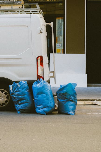 A small burgundy flatbed truck parked on a city street in front of a modern office building with large glass windows. The truck's cargo area is filled with various types of rubbish, including large wh